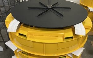 A close-up of a large, round yellow industrial part with foam padding at the edges—finished by an industrial powder coating—topped by a flat black circular component with spokes and a central rod, placed in a workshop or warehouse setting.