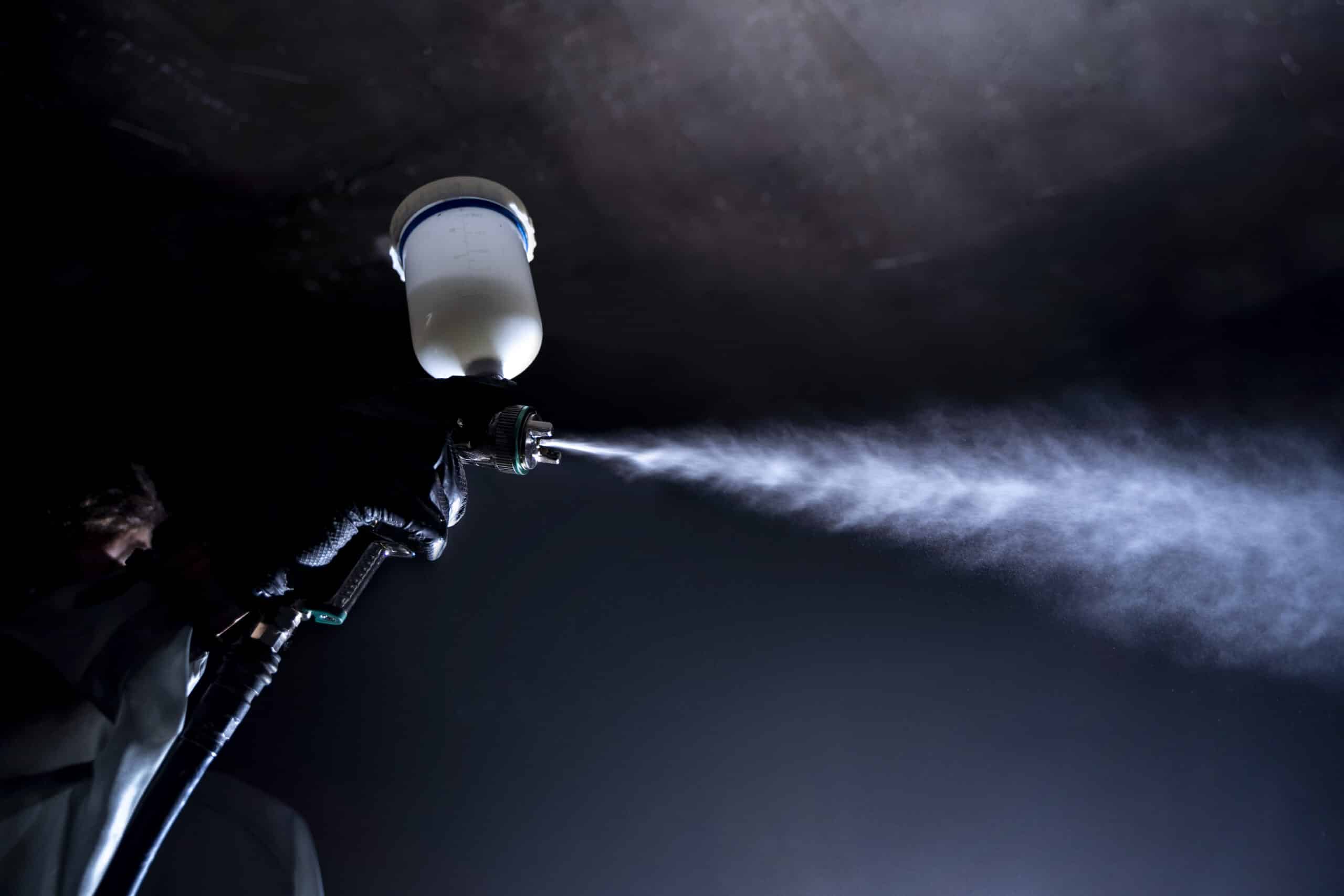 A person wearing black gloves uses a spray gun to emit a fine mist of paint, reflecting the precision OEMs in Minnesota & Wisconsin should look for in a powder coating partner, set against a dark background.